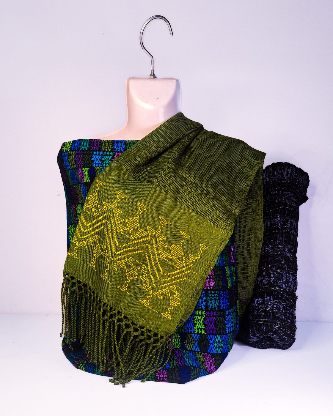 Conjunto con chal verde olivo bordado en amarillo, un textil de bloques de color y una pieza enrollada oscura