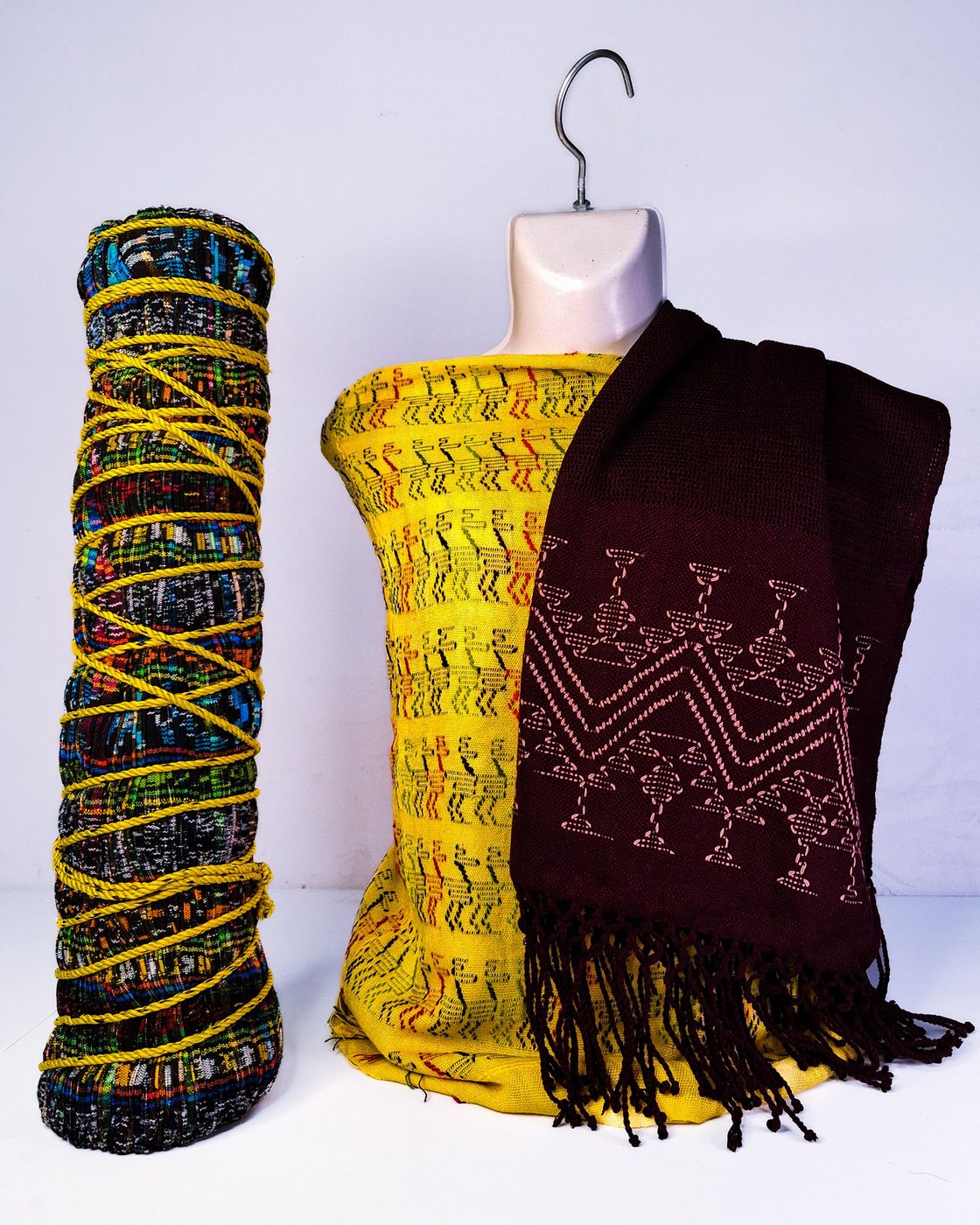 Conjunto con textil amarillo de figuras geométricas, chal café con bordado rosa y una pieza enrollada con cordón amarillo